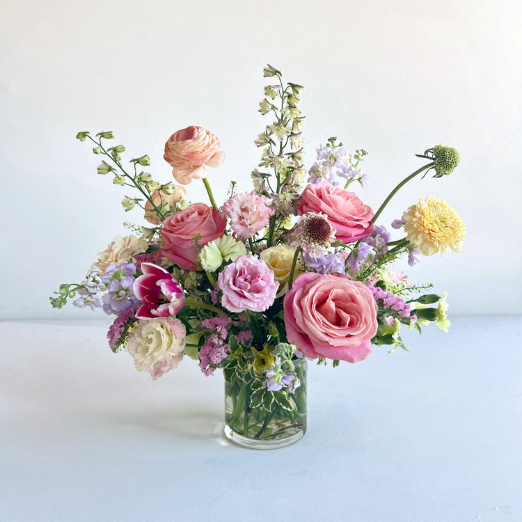 A beautifully arranged centerpiece of various flowers including roses and other foliage, in a clear glass vase placed on a white surface.